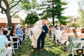 At a private residence in Watertown, Wisconsin, the groom joyfully carries his bride down the aisle following their heartfelt wedding vows outdoors in sunshine.